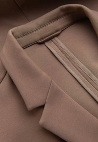 Brauner Stoff mit einer glatten Textur, ausgestattet mit einem umgeschlagenen Kragen und feinen Stickdetails entlang der Kanten. Einfaches und minimalistisches Design.