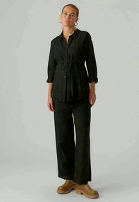 Črna srajca z gumbi in pasom, v kombinaciji s širokimi črnimi hlačami. Gladek material, minimalističen dizajn in svetlo rjave platforme.