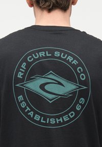 Άτομο που φοράει μαύρο μπλουζάκι με μπλε λογότυπο Rip Curl Surf Co και το κείμενο «Established 69» στην πλάτη.