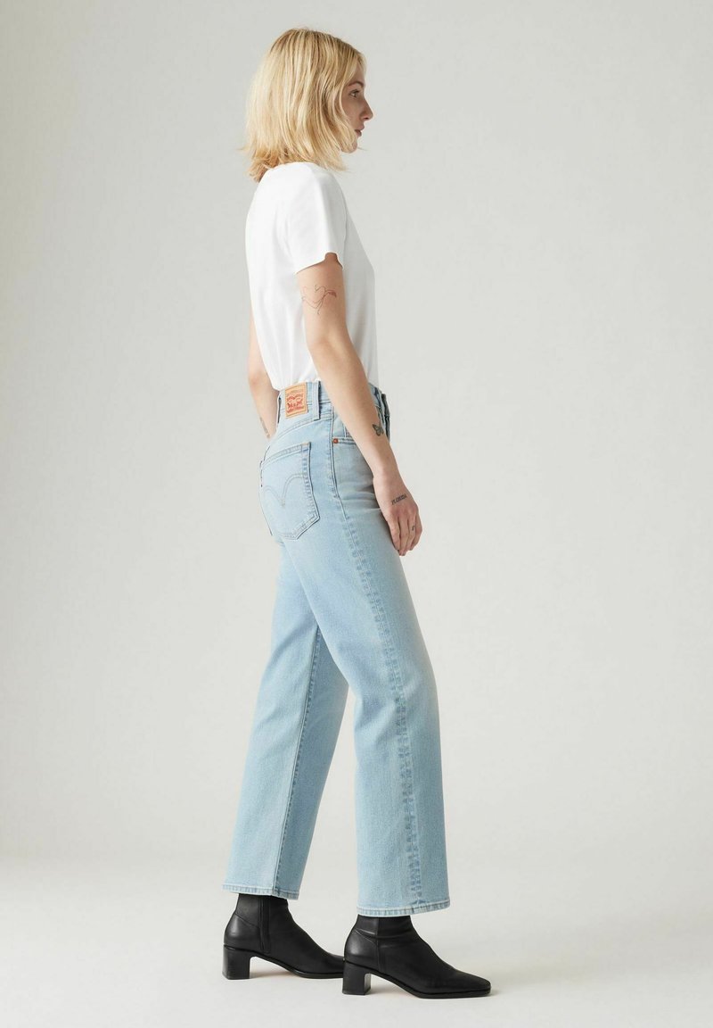 Camiseta blanca, jeans rectos de cintura alta en un tono azul claro y botas de tacón negro hasta el tobillo. Costuras de denim destacadas y diseño del bolsillo trasero.