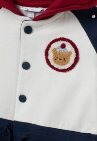 Sudadera crema y azul marino con un parche bordado circular de un oso con un sombrero de postre, asegurado con botones negros.