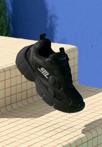 Black chunky sneaker with mesh design and "SNL" logo, positioned above a cream tiled surface against a blue wall background.