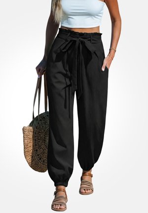 Vrouw draagt zwarte paperbag broek met hoge taille en strikceintuur, een wit cropped topje, sandalen en heeft een grote gewebde tas bij zich.