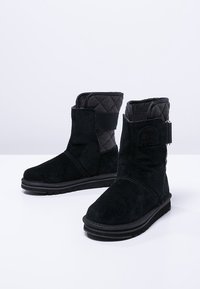 Schwarze Wildleder-Stiefeletten mit einem wattierten Innenfutter, runder Zehenform und einer flachen Gummisohle. Mit einem Klettverschlussriemen an der Seite.