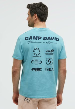 Man facing away wearing light blue Camp David t-shirt with sailing and surfing themed text and icons on the back.