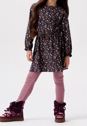 Jeune fille portant une robe longue à manches longues à motifs floraux avec une ceinture à la taille, des leggings roses côtelés et des bottes d'hiver violettes lacées, debout jambes écartées.