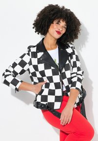 Veste en cuir synthétique à carreaux noir et blanc avec un col noir et des zips. Portée sur un t-shirt blanc et accessoirisée avec une ceinture noire.