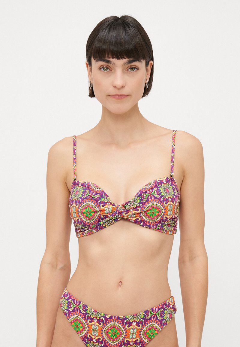 Vrouw met kort donker haar die een kleurrijke, gestreepte bikini draagt met paarse, groene en oranje geometrische bloemenpatronen.