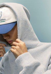 Svetlomodrá pletená mikina s textúrovaným vzorom, s veľkou kapucňou a rebrovanými manžetami. Nosí sa biela čiapka s modrými akcentmi.