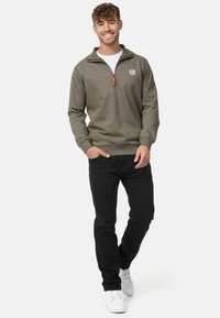 Grøn zip-up sweatshirt med et struktureret mønster, prydet med et hvidt logo og en brun lynlåsdetalje, parret med sorte bukser og hvide sneakers.