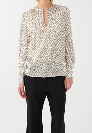 Kobieta w kremowej bluzce z czarnym wzorem w kropki i małym rozcięciem z przodu, w połączeniu z czarnymi spodniami, stojąca na białym tle.