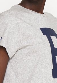 Grå bomullströja med korta ärmar som har ett stort marinblått bokstaven "F" tryckt på framsidan. Mjuk textur och rundad halsringning.