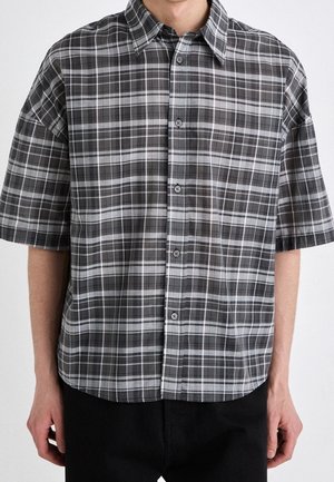 Homme portant une chemise à manches courtes à carreaux noirs et gris, se tenant devant un fond clair uni.
