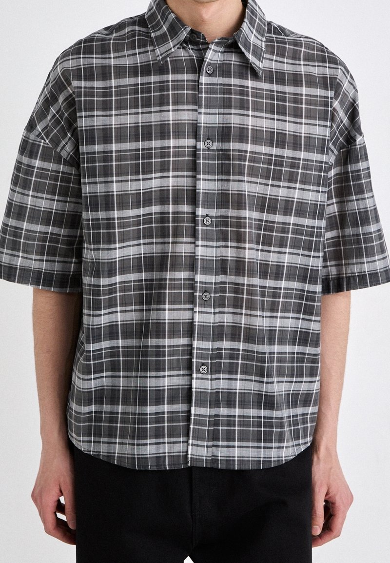 Homme portant une chemise à manches courtes à carreaux noirs et gris, se tenant devant un fond clair uni.