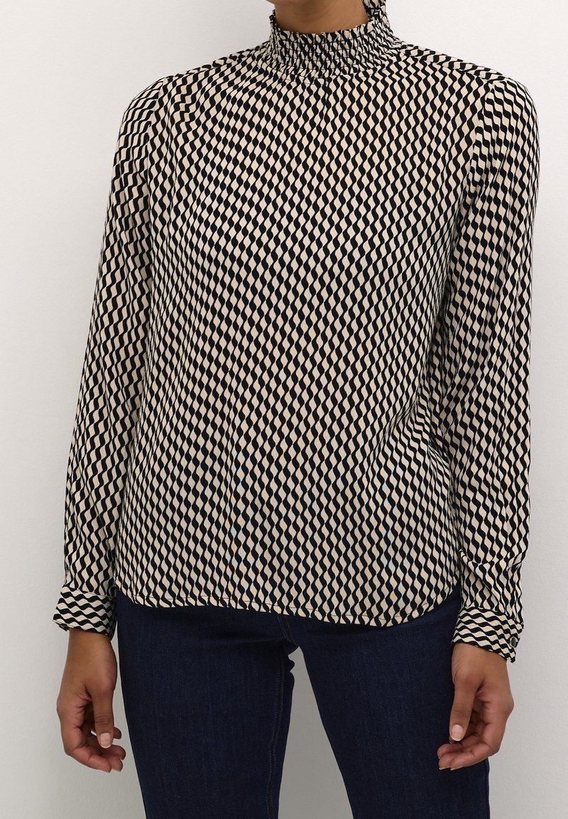 Langemouwenblouse met een hoge, geraffineerde halslijn. Heeft een geometrisch zwart-wit diamanten patroon op lichte stof.