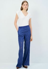 Camiseta blanca de cuello en V y mangas cortas, combinada con pantalones anchos de encaje azul con patrones intrincados y bolsillos. Unos zapatos marrones completan el atuendo.