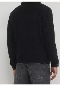 Homme portant une veste en velours côtelé noire avec des poignets boutonnés et un jean en denim noir délavé, vu de dos sur un fond uni.