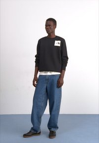 Svart hettegenser med en hvit logo på brystet, kombinert med løspassede blå jeans og mørke sko, satt mot en ensfarget bakgrunn.