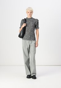Top preto com padrão e mangas bufantes, calças cinzentas de perna larga e bolsa preta. Modelo em pé sobre um fundo branco.