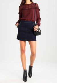 Donna che indossa un blouse in pizzo bordeaux, una mini gonna di jeans scuri con bottoni, stivali neri alla caviglia e una borsa a tracolla nera su uno sfondo bianco.