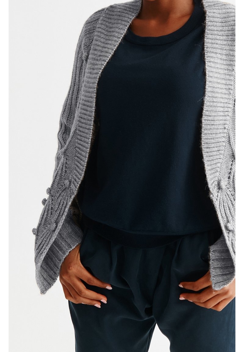 Personne portant un cardigan en maille gris ample sur un haut bleu foncé et un pantalon bleu foncé, les mains reposées sur les hanches.