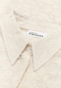 Koorene tekstuuriga kreemivärvi särk, millel on kortsus kangas, terava kraega ja nööbikinnitusega. Etiketil on kirjas "KAREN BY SIMONSEN."