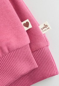 Dos mangas de tela tejidas en rosa con puños acanalados y etiquetas blancas; una etiqueta muestra un corazón marrón, la otra dice "smile little one."