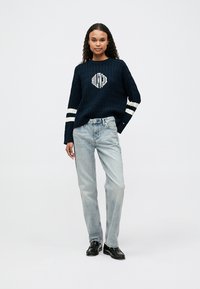 O tânără stă îmbrăcată cu un pulover bleu închis, tricotat în cablu, cu dungi albe, și blugi de un albastru deschis, purtând pantofi negri.