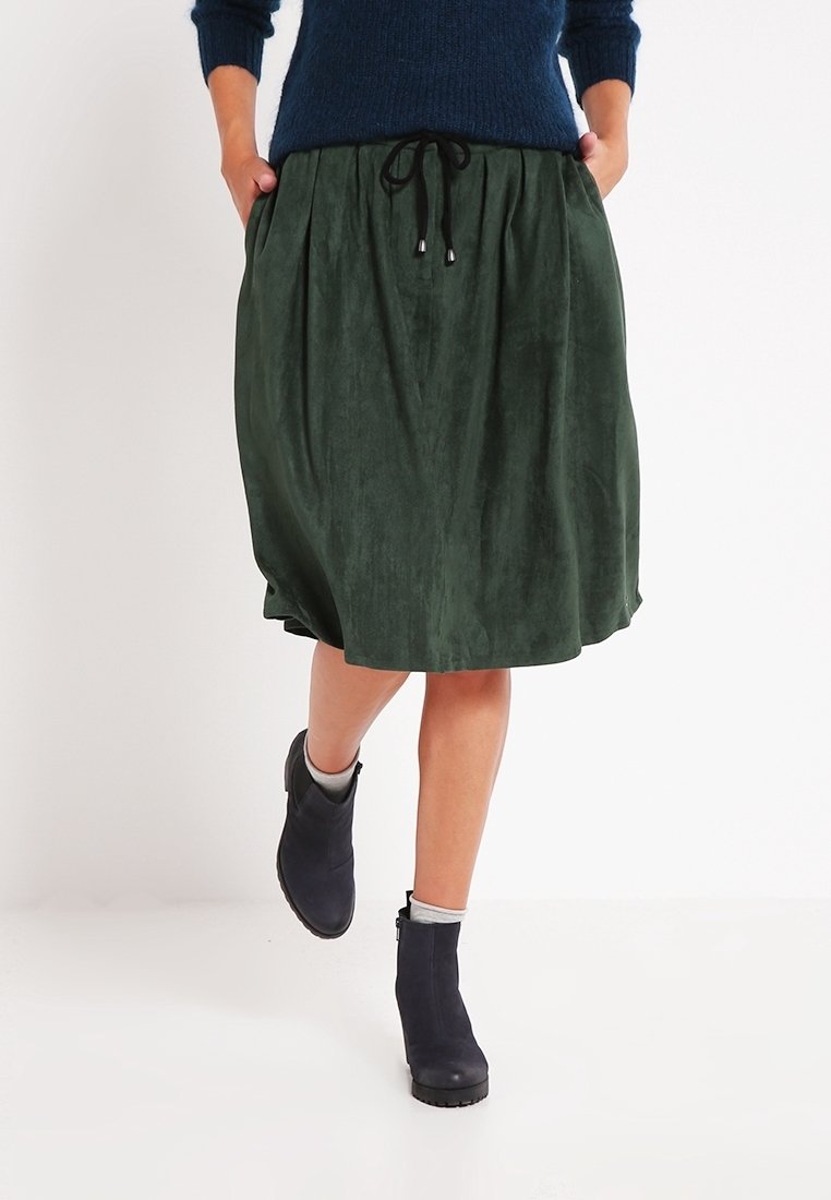 Falda de ante verde con pliegues, cintura con cordón ajustable y un corte relajado. Combinada con botas negras hasta el tobillo y un suéter de punto azul oscuro.