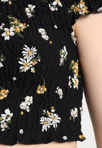 Černý top s květinovým vzorem a zmraženou texturou. Má bílé sedmikrásky a žluté akcenty, zakončené detailním lemováním.