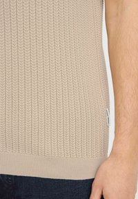Beige kötött pulóver, amely texturált halmintázattal, bordás szegéllyel és egy kis címkével rendelkezik az oldalon. Sötét anyag fölött viselve.