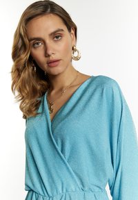 Šviesiai mėlyna aptraukimo suknelė su tekstūruotu, blizgančiu audiniu. Ilgos rankovės ir susirinkusi liemens dalis. Dėvima su auksiniais auskarais-žiedais ir sluoksniais kaklo papuošalų.