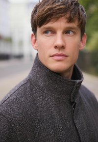 Ung man med blå ögon och kort brunt hår som b�är en mörkgrå knäppt rock, tittar åt sidan utomhus mot en suddig gatubakgrund.