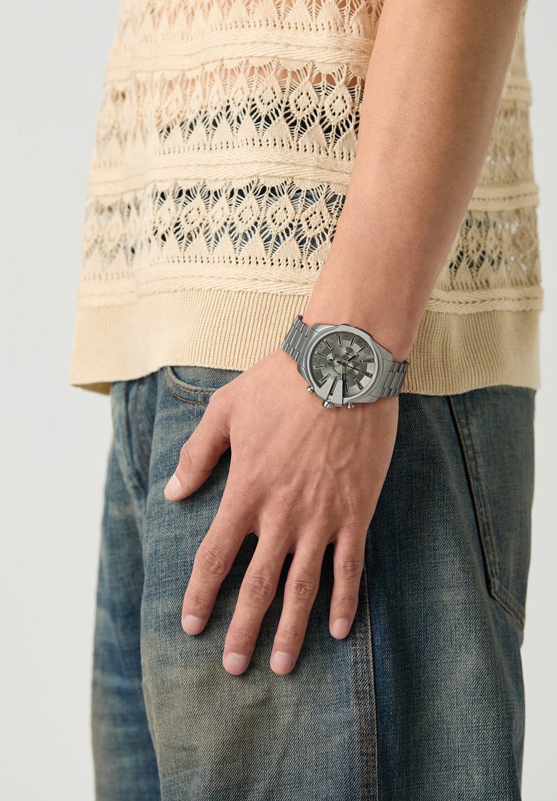 Mano che indossa un orologio in metallo argento con quadrante grande, appoggiata di lato; indossa un top lavorato a maglia color crema e jeans blu in denim.