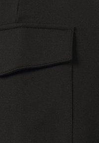 Černá tkanina s hladkým povrchem a mírným leskem, s detailem náprsní kapsy a kontrastním prošíváním podél okrajů.