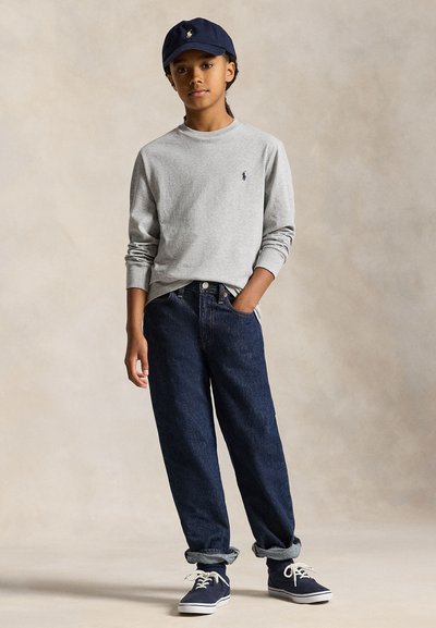 Mlada oseba, ki nosi temnomodro kapo, svetlo sivo majico z dolgimi rokavi, temne zavihane kavbojke in temno modre superge, stoji z eno roko v žepu.