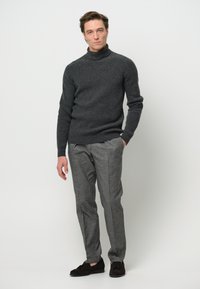 Grau gestrickter Rollkragenpullover kombiniert mit grauen maßgeschneiderten Hosen. Modell trägt dunkelbraune Wildlederschuhe. Schlichtes, strukturiertes und modernes Design.