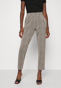 Calças em padrão houndstooth castanho e creme, com uma cintura alta e pernas ajustadas, usadas com uma blusa preta e sandálias de salto pretas.