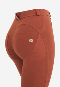 Leggings color ruggine con texture liscia, dotati di una vita alta, tasche curve e un'accenno di logo metallico sulla parte posteriore.