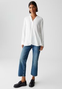 Vrouw met kort donker haar, gekleed in een losse witte blouse met lange mouwen, cropped blauwe jeans en zwarte veterschoenen, staande tegen een lichte achtergrond.