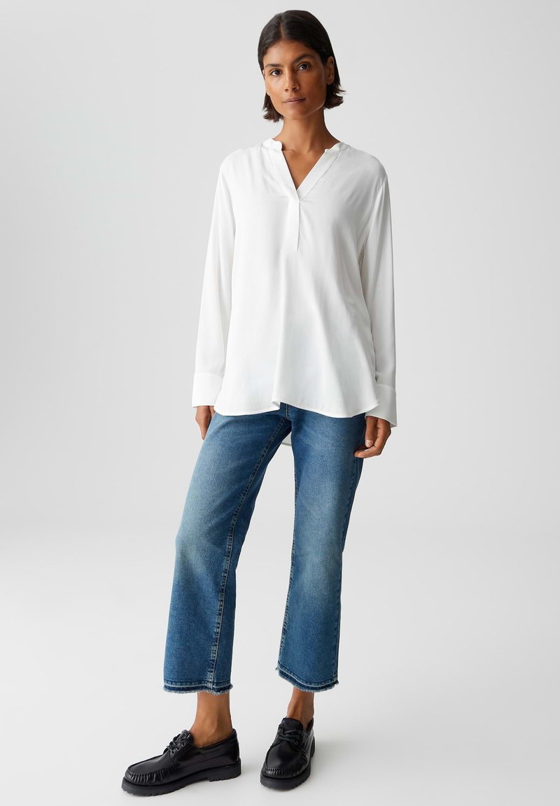 Vrouw met kort donker haar, gekleed in een losse witte blouse met lange mouwen, cropped blauwe jeans en zwarte veterschoenen, staande tegen een lichte achtergrond.