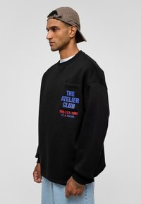 Zwarte sweatshort met een ronde hals, voorzien van een logo in blauw en rood op de borstzak. Gekocht met lichtblauwe jeans en een beige baseballcap.