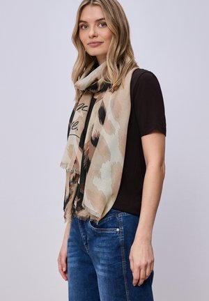 Kobieta o blond włosach, ubrana w czarną bluzkę z krótkim rękawem, niebieskie dżinsy i beżowy szal z wzorem, stojąca w pomieszczeniu na jasnoszarym tle.