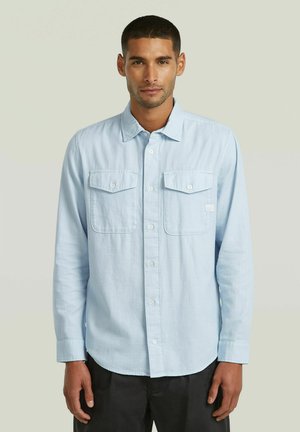 Mann mit kurzen Haaren, der ein hellblaues Hemd mit Knöpfen und zwei Brusttaschen trägt, steht vor einem einfarbigen hellen Hintergrund.