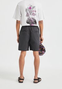 Shorts gris avec une taille élastique et deux poches arrière ; portés avec un t-shirt blanc arborant des motifs graphiques colorés au dos.