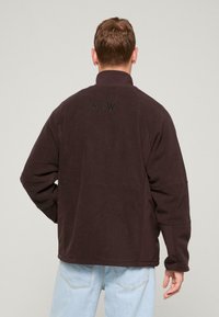 Mørkebrun fleecejakke med høj krave, der har de broderede bogstaver "ATW" på ryggen. Regular fit med lange ærmer.