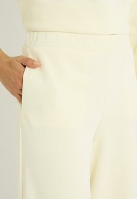 Tela acanalada de color crema, diseño de pierna ancha y bolsillo lateral. El material tiene una textura suave con un acabado delicado. Detalle visible en la pretina.