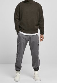 Maglione a collo alto texture verde scuro abbinato a pantaloni cargo grigi con tasche e polsini elasticizzati, completato da sneaker bianche.