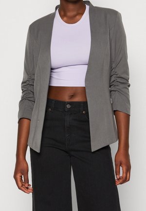 Person trägt einen grauen offenen Blazer, weißes kurzes Oberteil und hoch taillierte schwarze Hose, gezeigt vom Hals bis zu den Hüften.