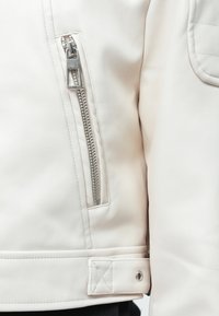 Valge nahktagi, millel on hõbedane tõmblukk rinna taskul, tikanditootnud muster õlavarre piirkonnas ja nupuga kinnitamine allservas.